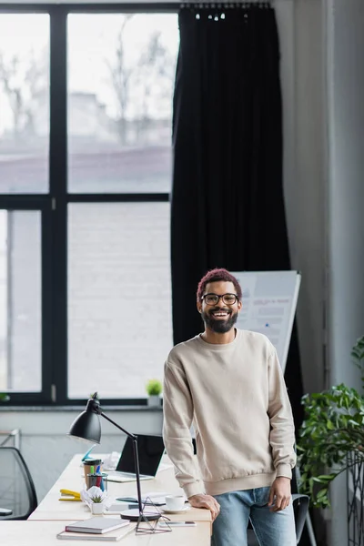 Joyeux homme d'affaires afro-américain à lunettes regardant la caméra près de la table de travail dans le bureau — Photo de stock