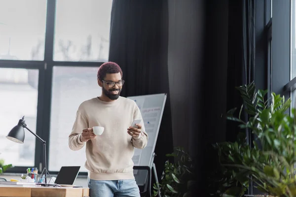 Felice uomo d'affari africano americano in possesso di tazza e utilizzando smartphone in caffè in ufficio — Foto stock