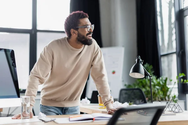 Empresário afro-americano sorridente que está perto da água e do computador no escritório — Fotografia de Stock