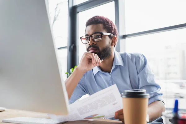 Homme d'affaires afro-américain concentré dans les lunettes et les vêtements formels tenant du papier près de l'ordinateur dans le bureau — Photo de stock