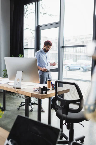 African american businessman holding paper and smartphone near coffee and computer in office — Stock Photo