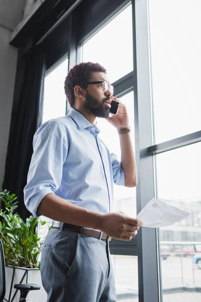 Low angle view of african american businessman in eyeglasses talking on smartphone and holding document in office — Stock Photo