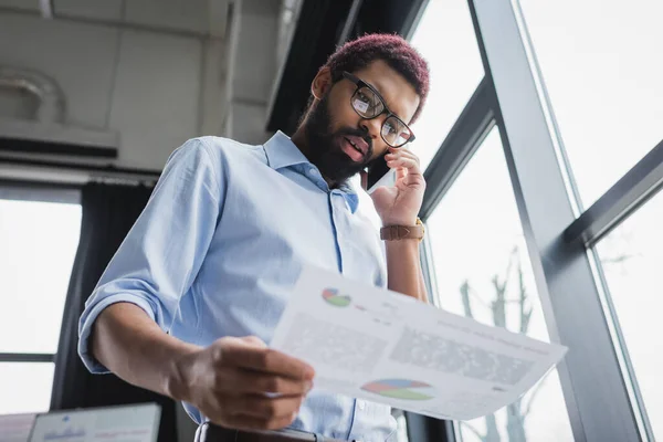 Low angle view of african american businessman talking on smartphone and holding paper in office — Stock Photo