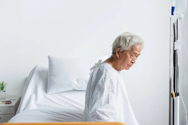 Triste paciente asiático sentado en la cama cerca de la terapia intravenosa en la sala de hospital - foto de stock