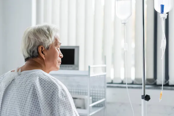 Vista lateral del paciente asiático de edad avanzada que mira la terapia intravenosa en el hospital - foto de stock