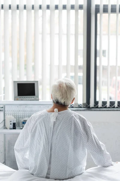 Back view of senior patient in gown sitting on bed in hospital ward — Stock Photo