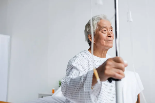 Elderly asian patient holding blurred stand with intravenous therapy in hospital ward — Stock Photo