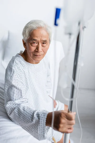 Senior asiático paciente mirando cámara cerca borrosa stand con intravenoso terapia en clínica — Stock Photo