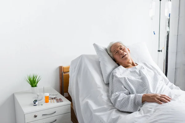 Ill asiático paciente deitado na cama perto de pílulas e água no hospital ward — Fotografia de Stock