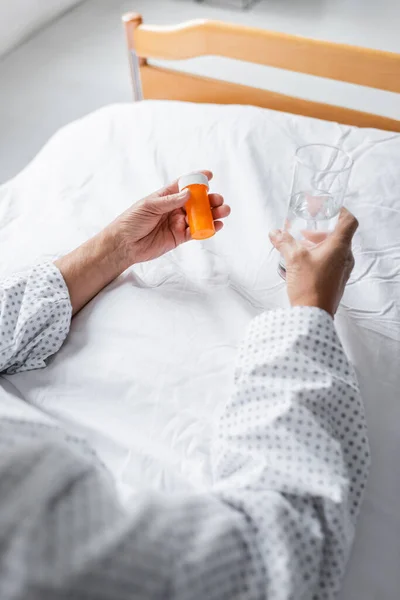 Cropped view of senior patient holding water and pills on bed in clinic — Stock Photo