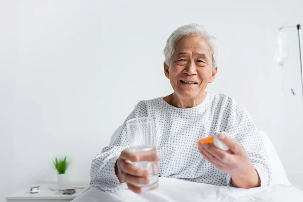 Веселий азіатський пацієнт тримає розмиті склянки води і таблетки на ліжку в лікарняному палаті — стокове фото