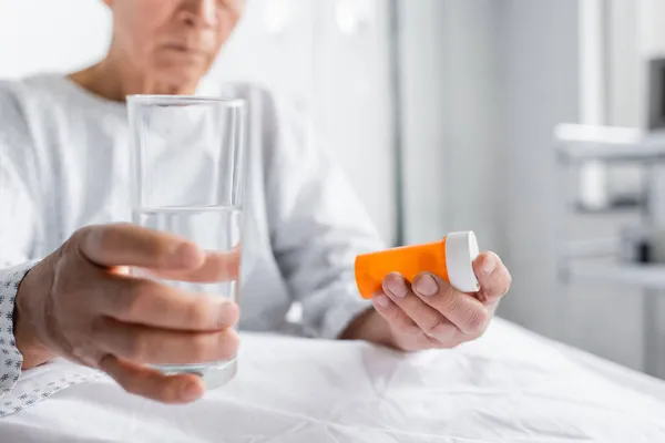 Vista cortada de pílulas e água nas mãos do paciente desfocado na enfermaria do hospital — Stock Photo