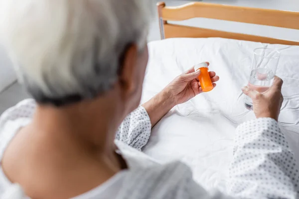 Blurred elderly patient holding pills and glass of water on bed in hospital — Stock Photo