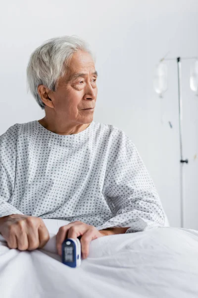 Літній азіатський пацієнт з пульсоксиметром сидить на ліжку в лікарняному палаті — стокове фото