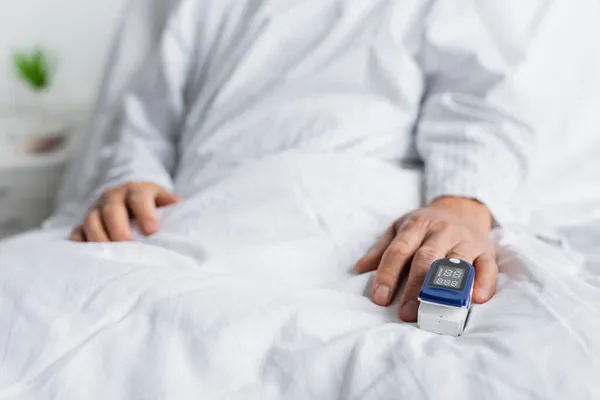 Vista recortada del oxímetro de pulso en la mano del paciente anciano borroso en la cama en la clínica - foto de stock