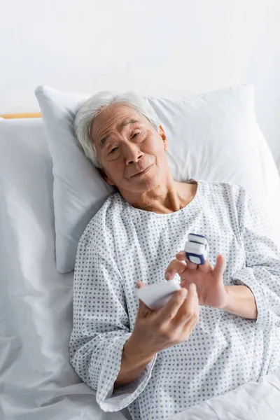 Paciente asiático de edad avanzada con oxímetro de pulso usando smartphone en la sala de hospital - foto de stock
