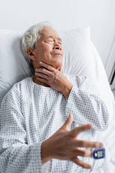 Enfermo asiático paciente con pulso oxímetro tocando cuello en cama en clínica - foto de stock