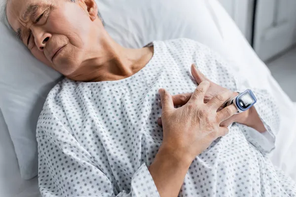 Vista de ángulo alto del paciente asiático enfermo con oxímetro de pulso acostado en la cama en la clínica - foto de stock