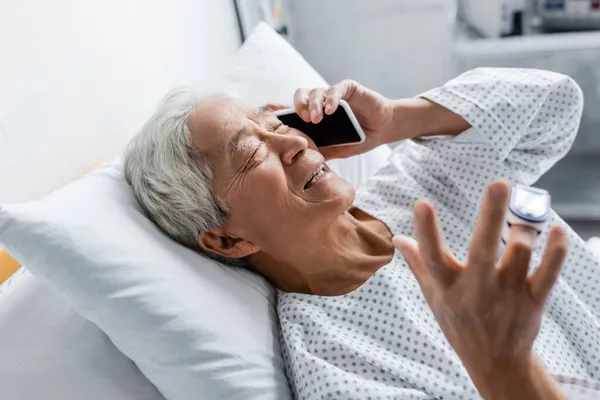 Sonriente paciente asiático con oxímetro de pulso hablando en smartphone en clínica - foto de stock