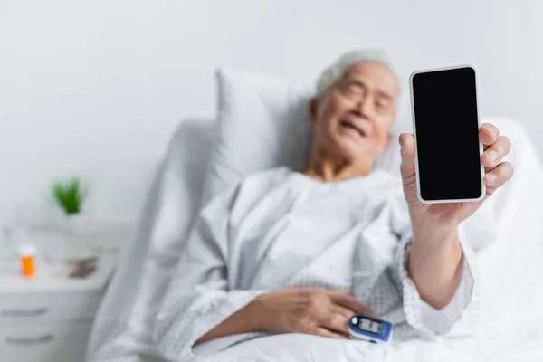 Smartphone in mano del paziente asiatico offuscato con pulsossimetro nel reparto ospedaliero — Foto stock