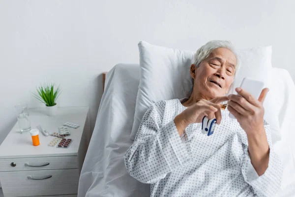 Patient asiatique âgé avec oxymètre de pouls utilisant un smartphone sur le lit près des pilules et de l'eau à la clinique — Photo de stock
