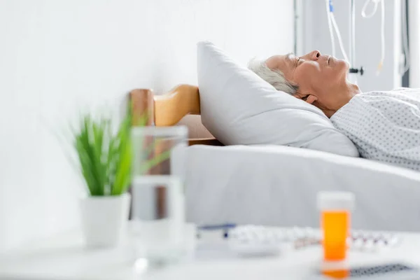 Vista lateral de paciente asiática mayor durmiendo en la cama cerca de agua borrosa y pastillas en la clínica — Stock Photo