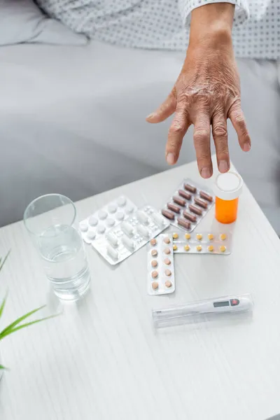 Vista recortada del paciente mayor tomando píldoras cerca del termómetro y el agua en la sala de hospital - foto de stock