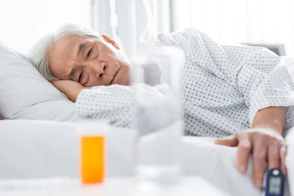 Paciente asiático de edad avanzada con oxímetro de pulso mirando píldoras y agua en la clínica - foto de stock