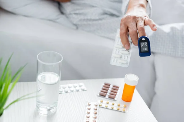 Ausgeschnittene Ansicht eines älteren Patienten mit Pulsoximeter, der Tabletten in der Nähe eines Wasserglases auf der Krankenhausstation einnimmt — Stockfoto