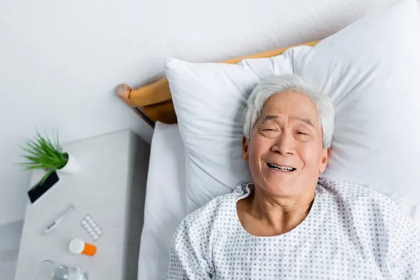 Draufsicht auf fröhliche asiatische Patientin, die auf Krankenhausstation im Bett liegt — Stockfoto