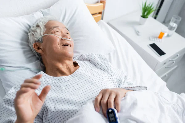 Vue grand angle du patient asiatique souriant avec oxymètre de pouls et canule nasale couchée sur le lit en clinique — Photo de stock