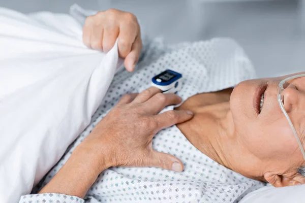 Vista recortada del anciano enfermo con cánula nasal y oxímetro de pulso acostado en la cama en la clínica - foto de stock