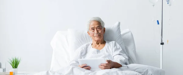 Sênior asiático paciente com cânula nasal segurando comprimido digital perto de terapia intravenosa na enfermaria do hospital, banner — Fotografia de Stock