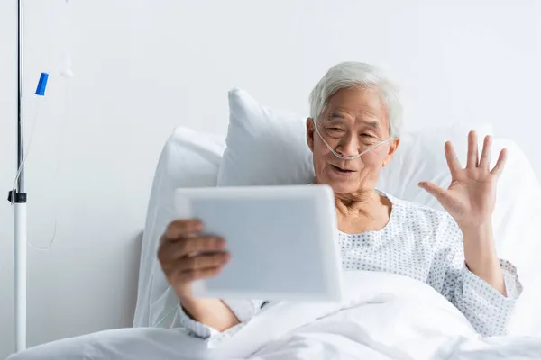 Старший азиатский пациент с носовой канюлей, имеющий видеозвонок на цифровом планшете в клинике — стоковое фото
