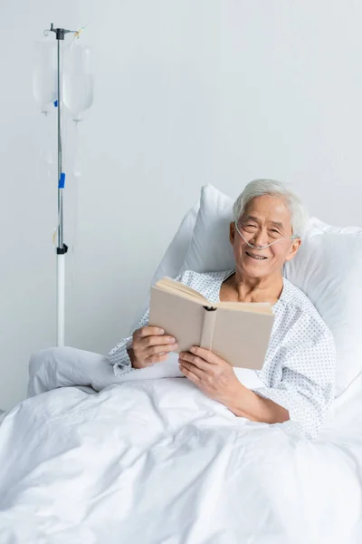 Sonriente paciente asiático con cánula nasal sosteniendo libro cerca de la terapia intravenosa en la sala de hospital - foto de stock