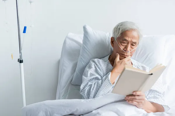 Elderly asian patient with nasal cannula reading book on bed in clinic — Stock Photo