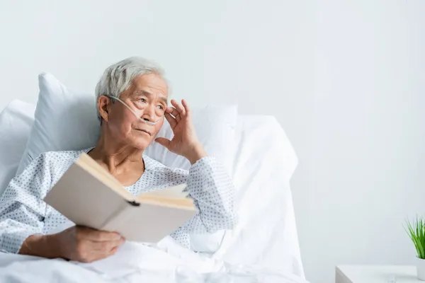 Asiática paciente en vestido y cánula nasal celebración libro en la cama en la clínica - foto de stock