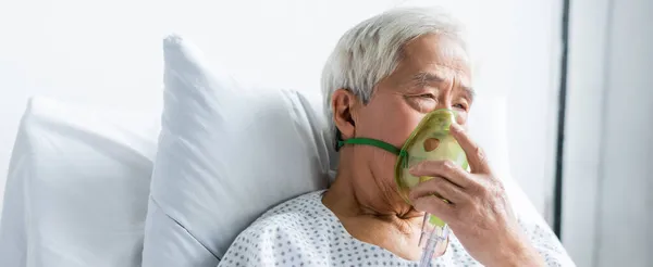 Старший азіатський пацієнт у кисневій масці сидить на ліжку в клініці, банер — стокове фото