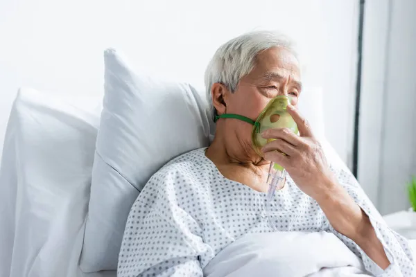 Senior asian patient holding oxygen mask while sitting on bed in hospital — Stock Photo