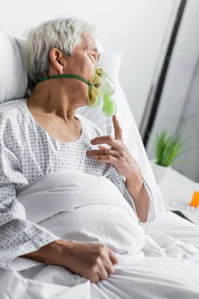 Vue latérale du patient asiatique en masque à oxygène couché sur le lit dans la salle d'hôpital — Photo de stock