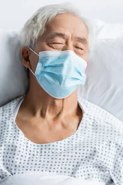 Senior asian patient in medical mask and gown lying on bed in clinic — Stock Photo