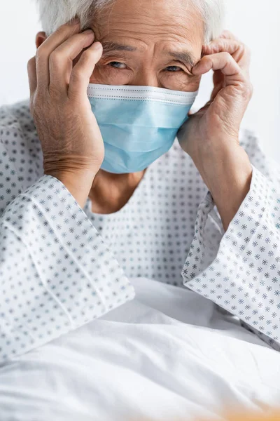 Enfermo asiático paciente en médico máscara mirando cámara en cama en hospital - foto de stock