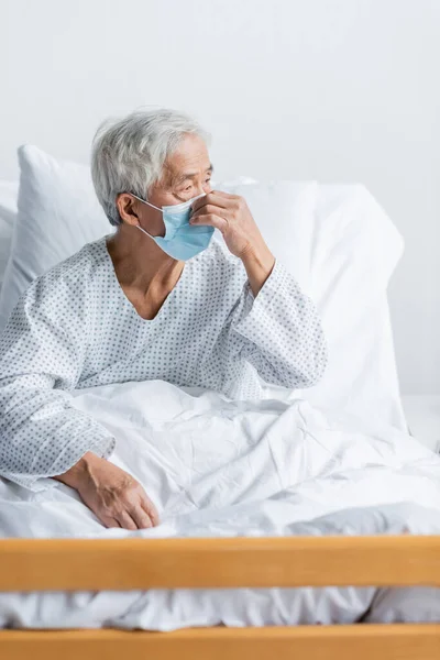 Ältere asiatische Patientin in medizinischer Maske sitzt auf Krankenhausbett — Stockfoto