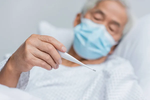 Offuscata asiatico paziente in medico maschera holding termometro su letto in clinica — Foto stock