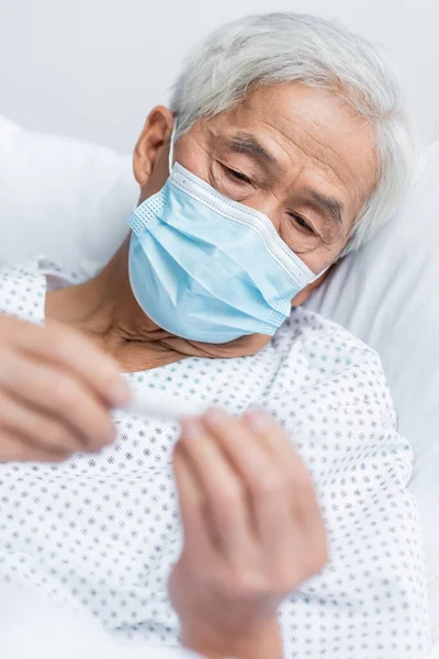 Senior asian patient in medical mask holding blurred thermometer on bed — Stock Photo