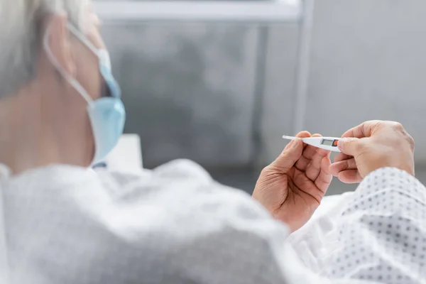 Verschwommene Seniorin mit Maske hält Thermometer auf Krankenhausstation — Stockfoto