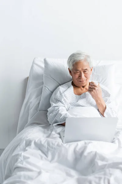 Старший азіатський пацієнт тримає склянку води і дивиться на ноутбук на ліжку в клініці — стокове фото