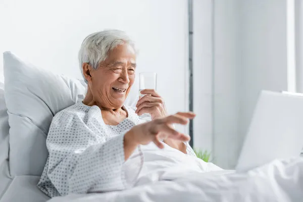 Cheerful asian patient with glass of water looking at laptop on bed in hospital — Stock Photo