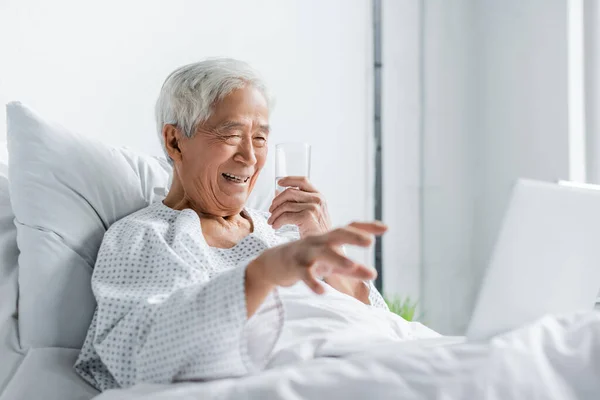 Cheerful asian patient with glass of water looking at laptop on bed in hospital — Stock Photo