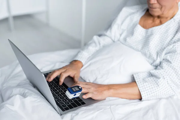 Cropped view of blurred patient in gown and pulse oximeter using laptop on hospital bed — Stock Photo